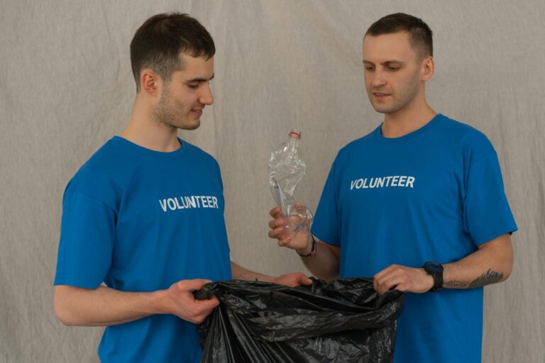 Two male volunteers in blue shirts collecting plastic bottles for recycling indoors.